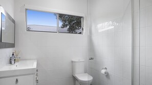 A bathroom with white tiles, a toilet, a sink with a mirror, and a window above the toilet. - Heated swimming pool and walk to beach (BATEAU BAY)