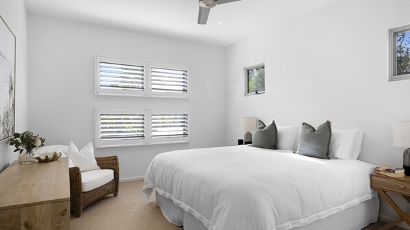 A bedroom with a large bed, two windows with blinds, a desk with a chair, and a nightstand with a lamp.