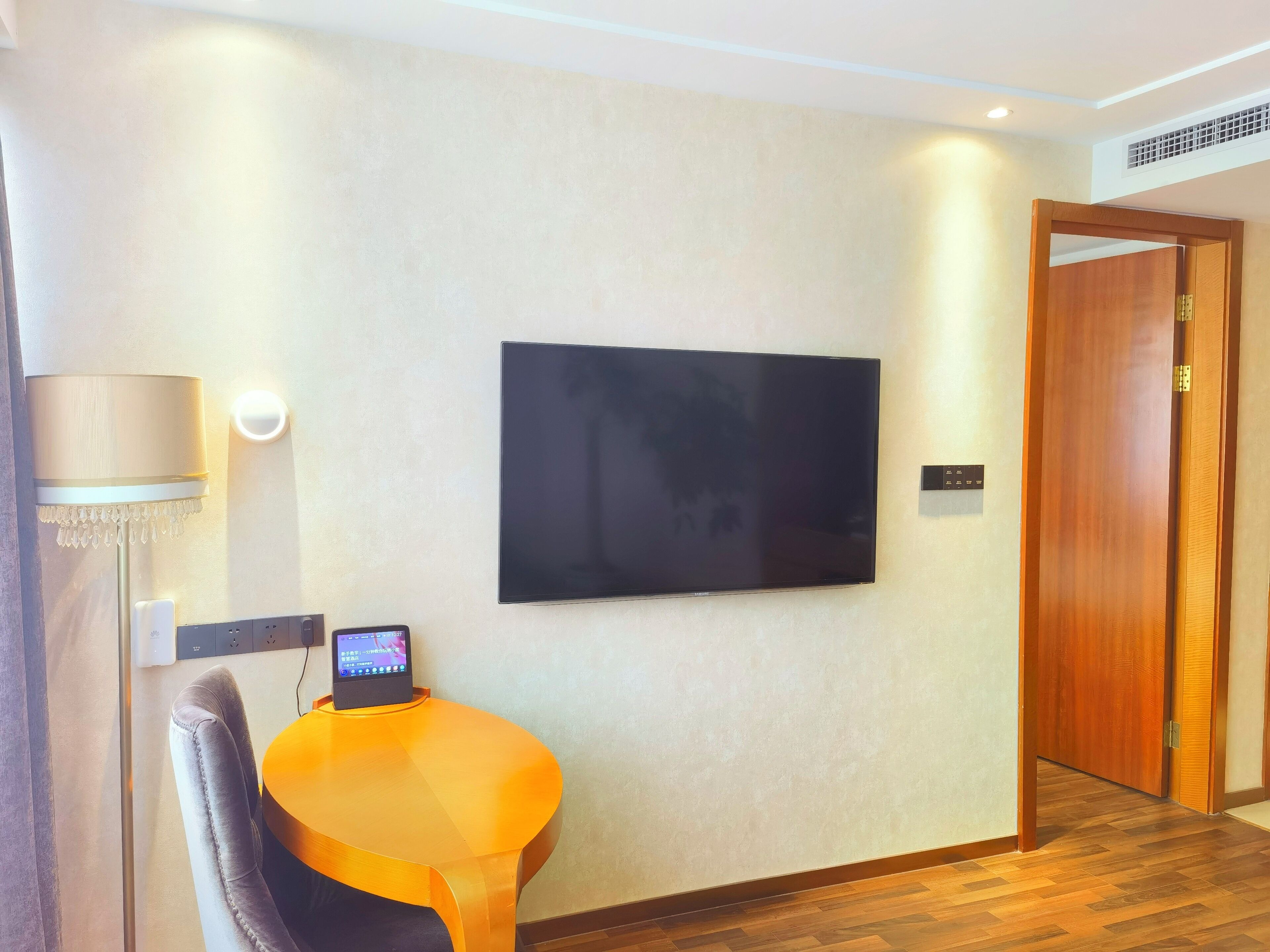 A hotel room with a flat-screen TV mounted on the wall, a small round table with a chair, a lamp, and a wooden door.