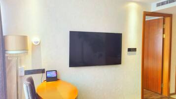 A hotel room with a flat-screen TV mounted on the wall, a small round table with a chair, a lamp, and a wooden door.