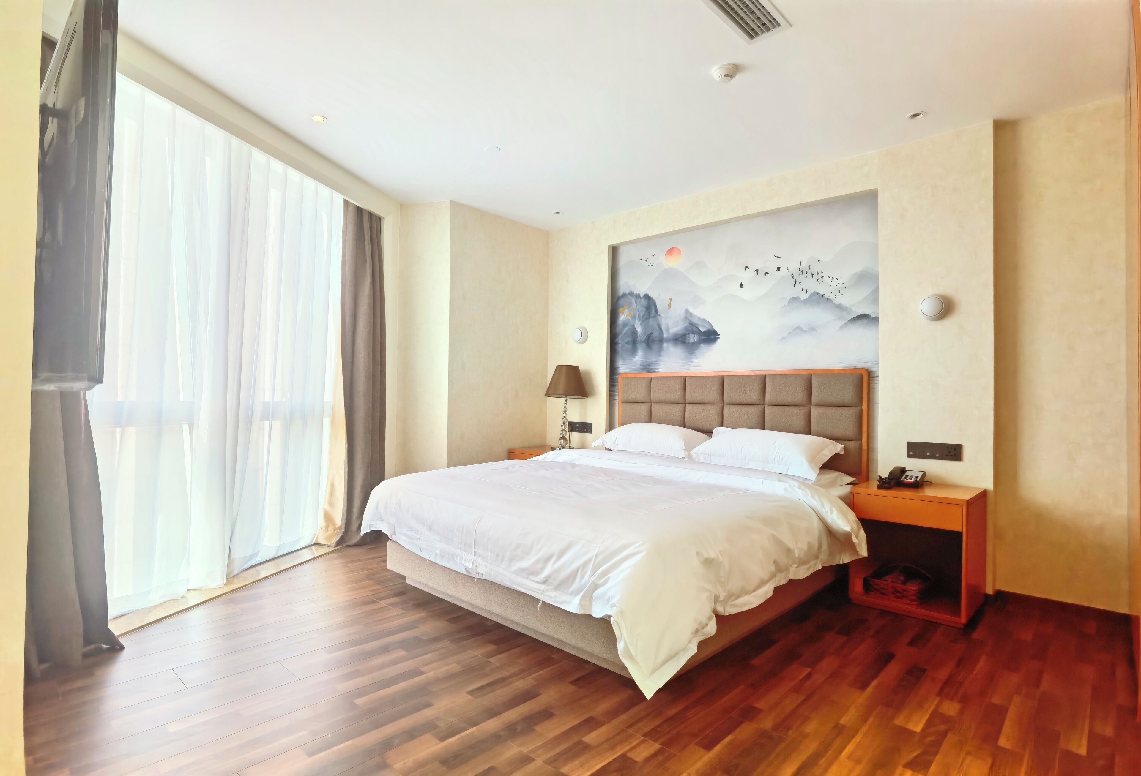 A spacious bedroom with a large bed, wooden flooring, a nightstand with a lamp, and a wall-mounted artwork.