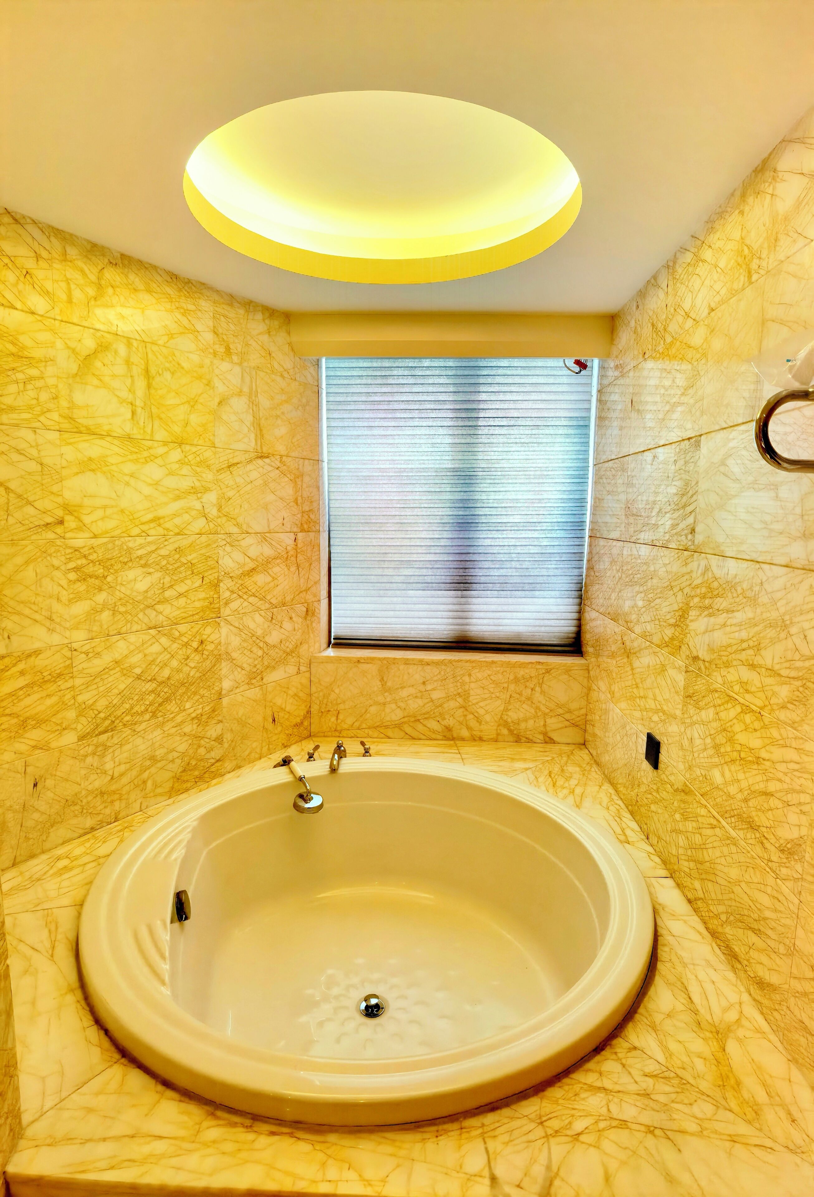A bathroom with a marble wall, a bathtub, a window with blinds, and a ceiling light.
