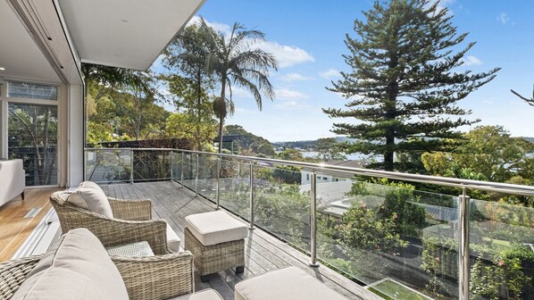 A balcony with wicker furniture, glass railings, and a view of trees and a lake. - Beautiful views, beachs and bushwalks (WAGSTAFFE)