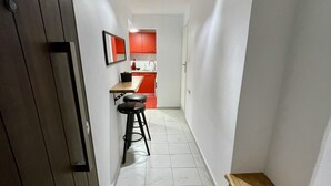 A narrow hallway with a wooden bench, a stool, and a door leading to a kitchen with red cabinets. - Central Studio Flat with Garden (Volos)