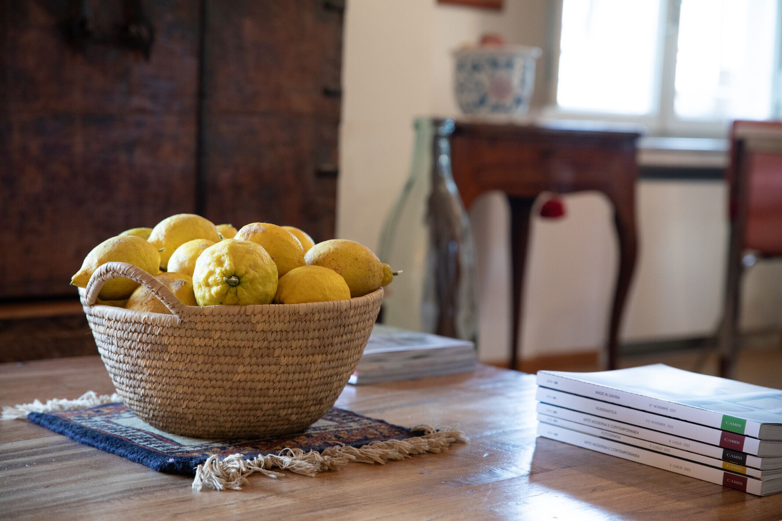 A woven basket filled with lemons on a wooden table, with a stack of books beside it.
