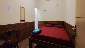 A small, single-bed room with a wooden headboard, a red bedspread, a wooden chair, a small table, and a brick accent wall. - Hotel O Aksana Homestay SyariahNearWonderland Trik Art (Purwokerto)