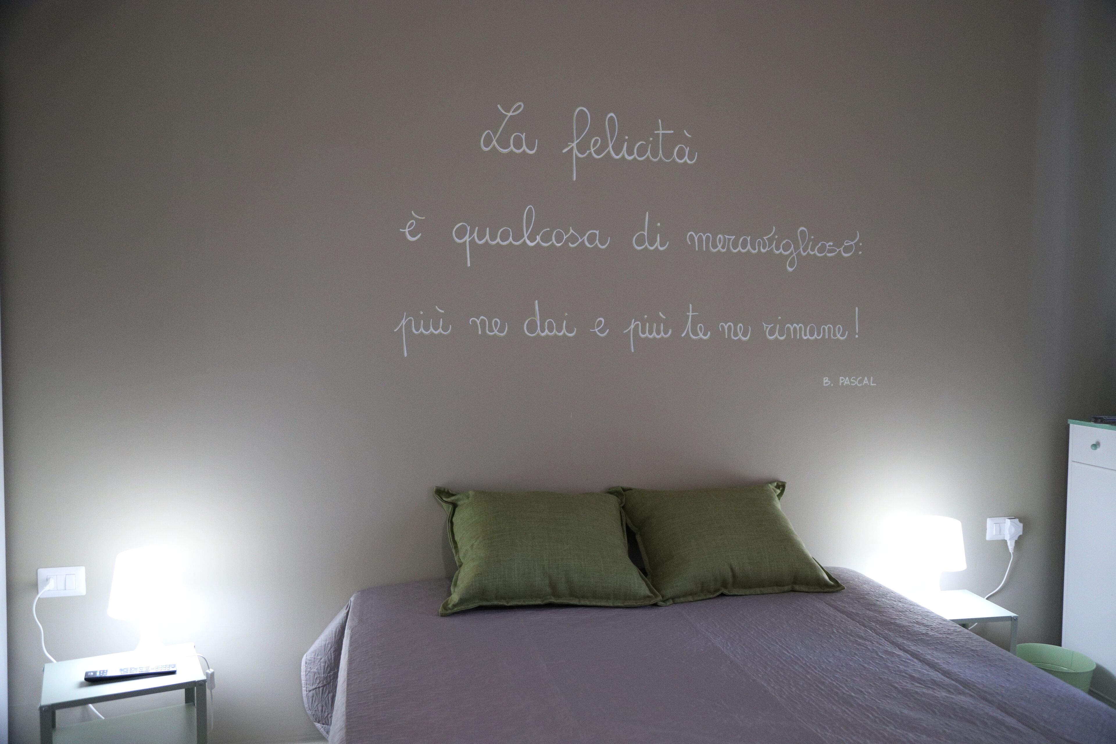 Een slaapkamer met een bed, kussens, een nachtkastje, een lamp en een muur met een citaat in het Italiaans.