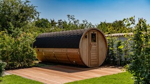 Een houten, vatvormige constructie met een deur, omgeven door een houten terras en weelderige begroeiing.