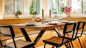 Een eettafel gedekt met borden, glazen en kaarsen, omringd door rieten stoelen, met uitzicht op groen door grote ramen.