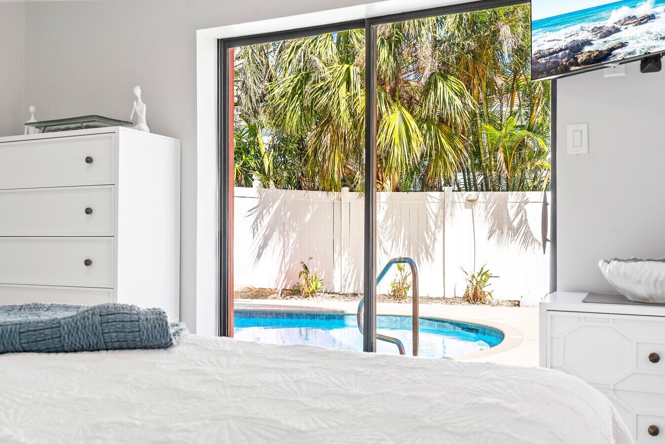 A bedroom with a bed, a dresser, and a view of a swimming pool and palm trees.