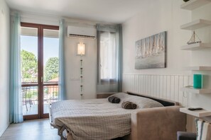 A bedroom with a bed, a bedside table, a window with curtains, a wall-mounted air conditioner, and a view of the outdoors.
