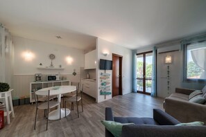 Ein modernes Wohnzimmer mit einer Couch, einem Esstisch mit Stühlen, einer Küchenzeile inklusive Mikrowelle und einer Schiebeglastür, die zu einem Außenbereich führt.