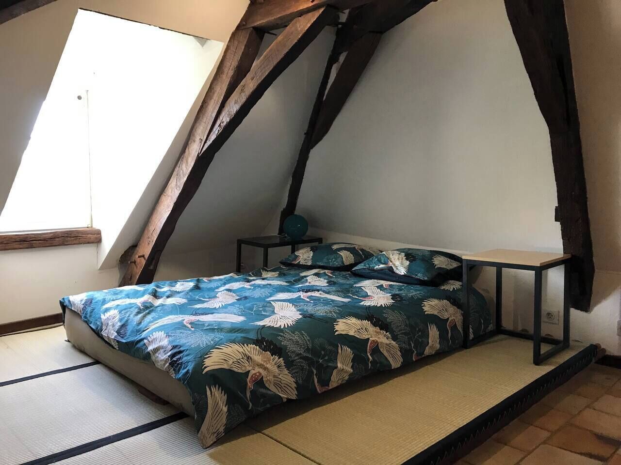 A small, cozy room with a sloped ceiling, a bed with a floral bedspread, a small table, and a window providing natural light.