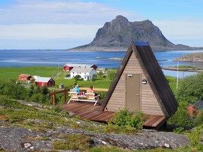 A wooden triangular cabin with a door and a small window, situated on a wooden deck with two chairs. - BaseCamp Vega (Vega)
