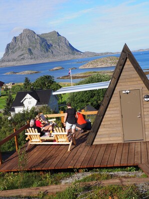 A wooden deck with a view of a mountain and sea, a small building with a door, and a flag. - BaseCamp Vega (Vega)