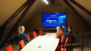 Une salle de conférence avec une longue table et des chaises rouges, un grand écran diffusant une présentation, et un plafond en pente avec des poutres en bois.
