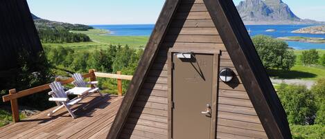 A wooden triangular cabin with a door marked "18" and a mailbox. It has a deck with two white chairs and a view of a lake and mountains.