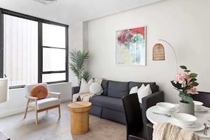 A modern living room with a grey sofa, a wooden chair, a round side table, and a dining area with white plates and bowls.