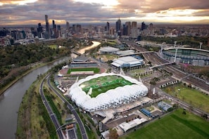 An aerial view of a stadium with a unique geodesic dome roof, surrounded by a river, roads, and a city skyline in the background.