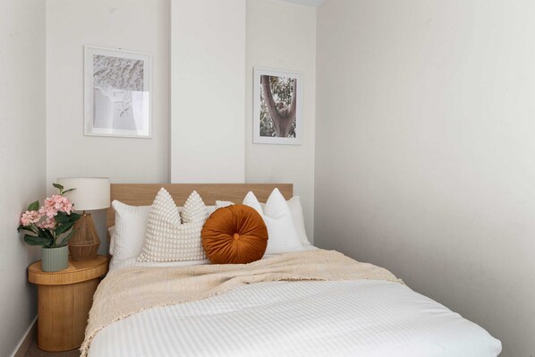 Un dormitorio con cama, almohadas, una mesita de noche con lámpara y flores, y dos cuadros en la pared.