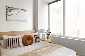 A neatly arranged bedroom with a bed, pillows, a bedside table with a lamp, a vase with flowers, and a window with a view of the sea.