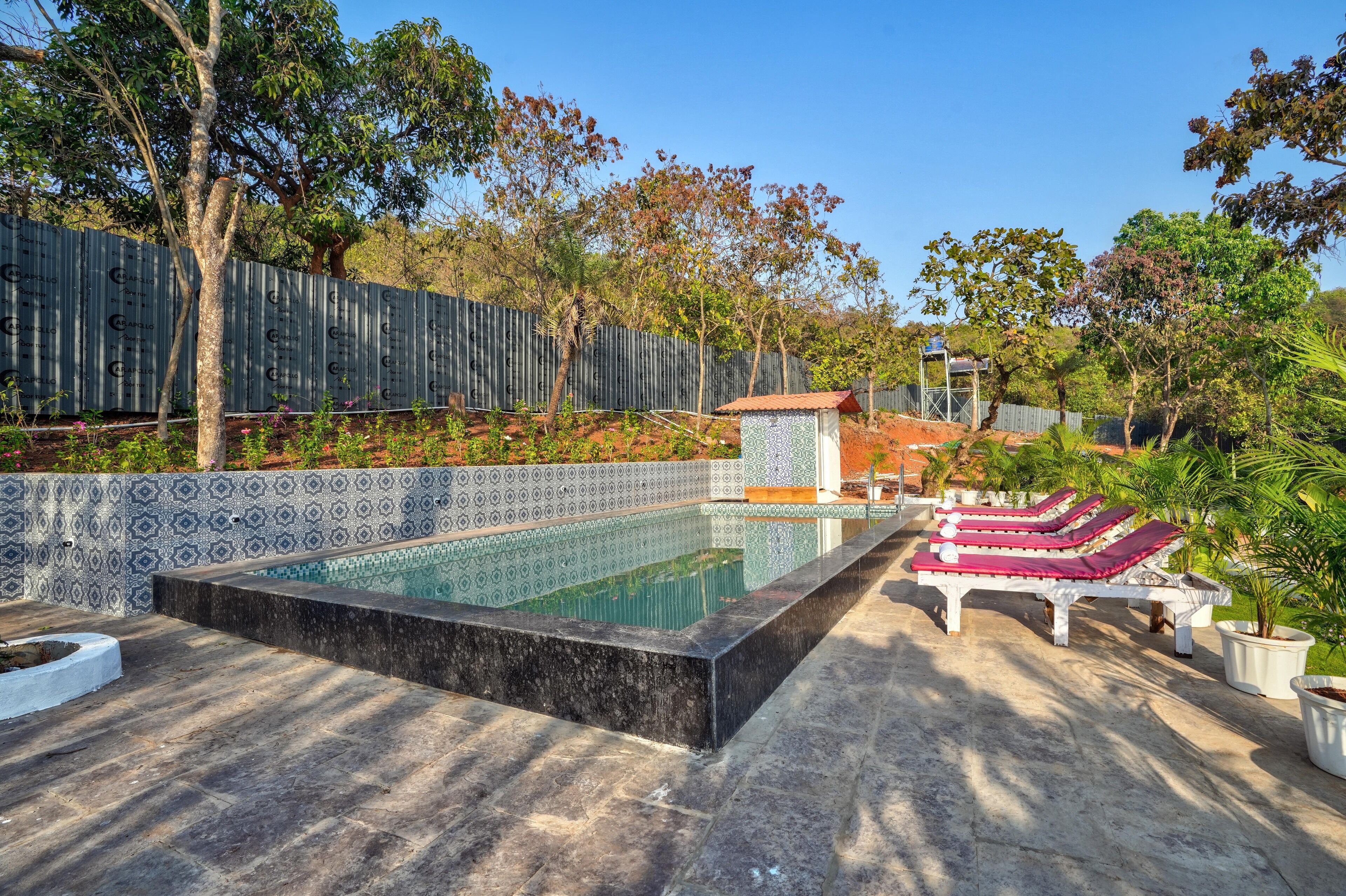 A swimming pool with a tiled edge, surrounded by a patterned wall and a fence. There are lounge chairs with red cushions and a small structure with a tiled roof.