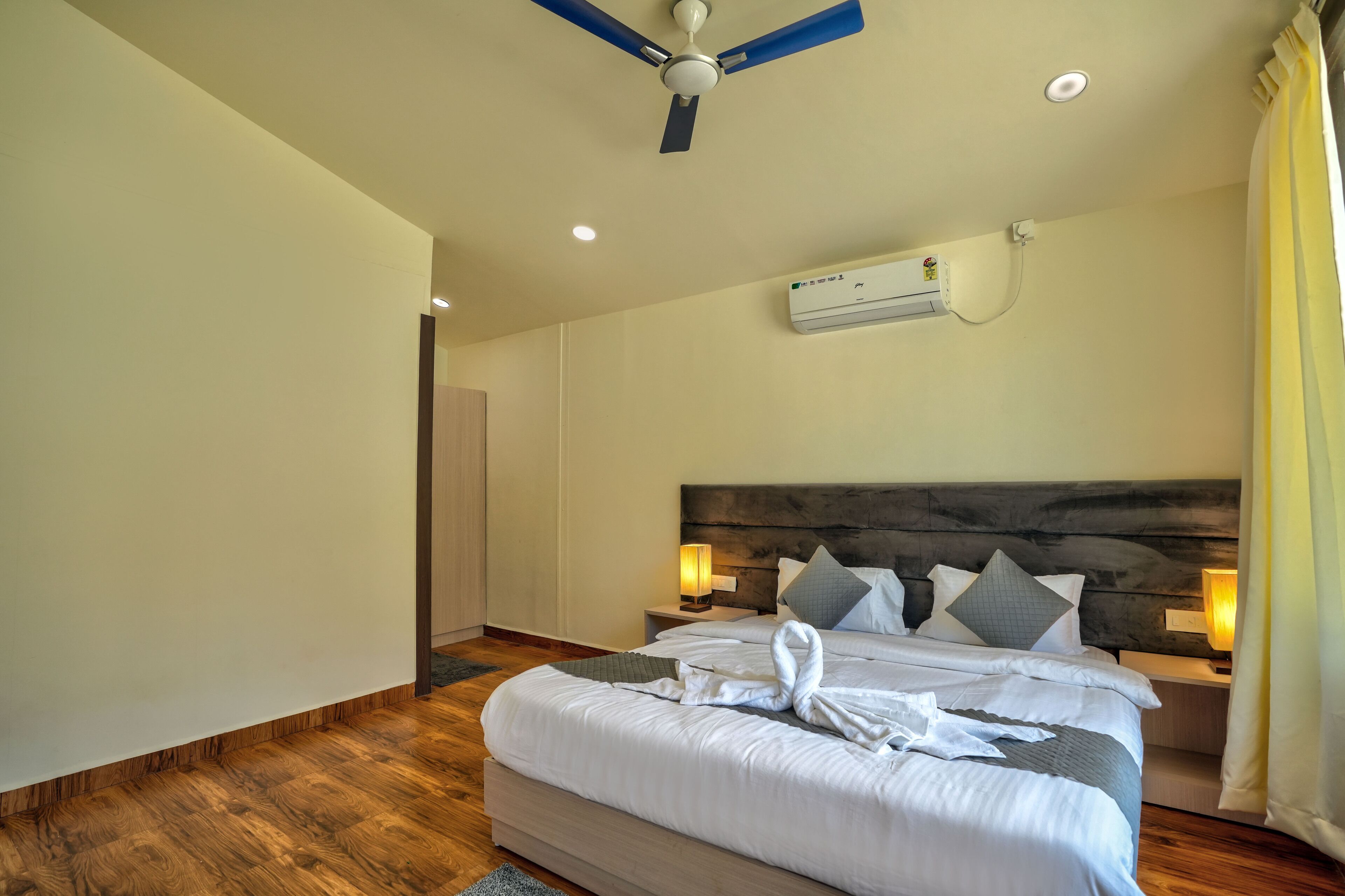A hotel room with a large bed, a ceiling fan, an air conditioning unit, and a nightstand with a lamp.