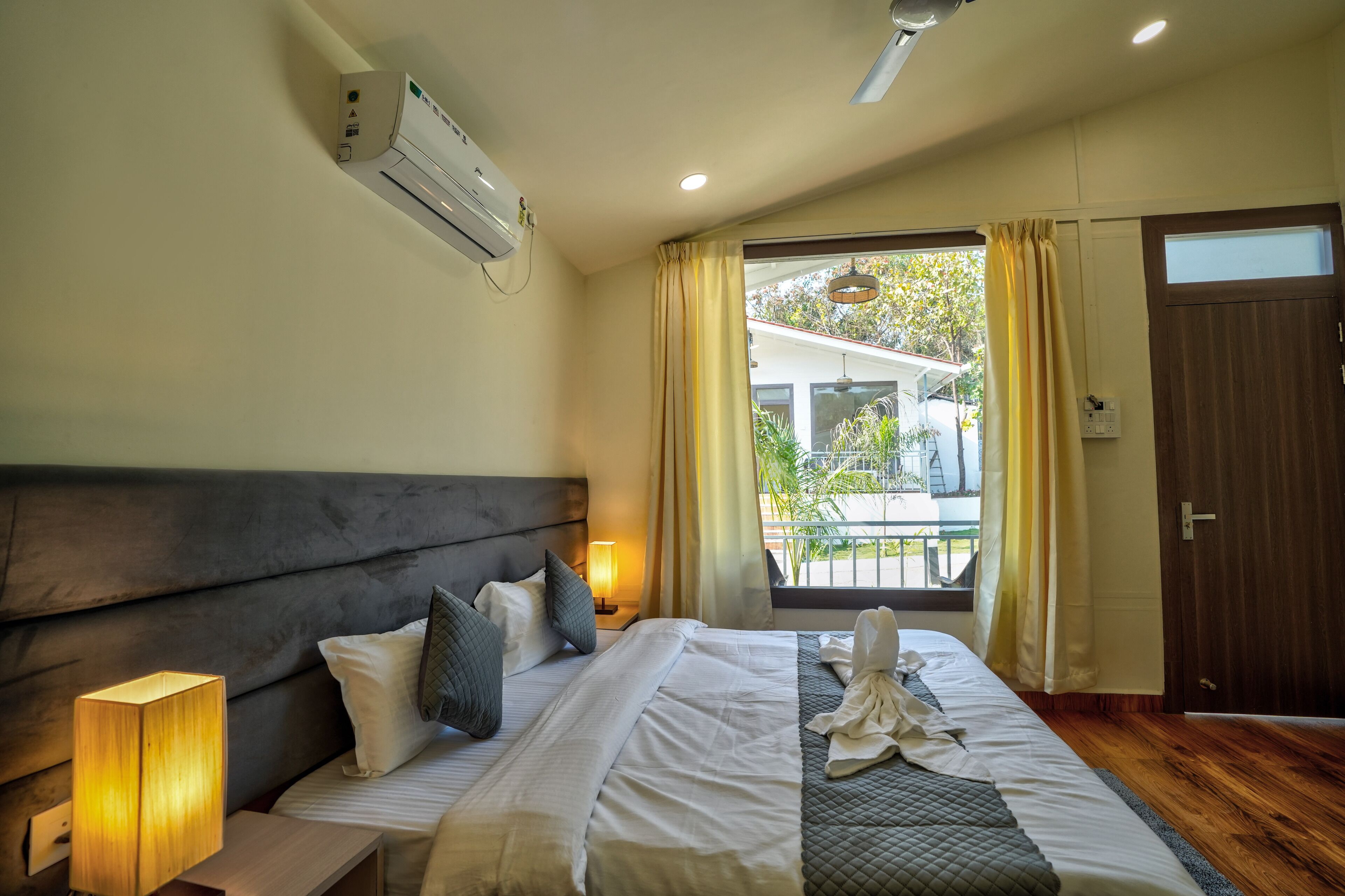 A hotel room with a bed, bedside lamp, air conditioning unit, and a view of the outside.