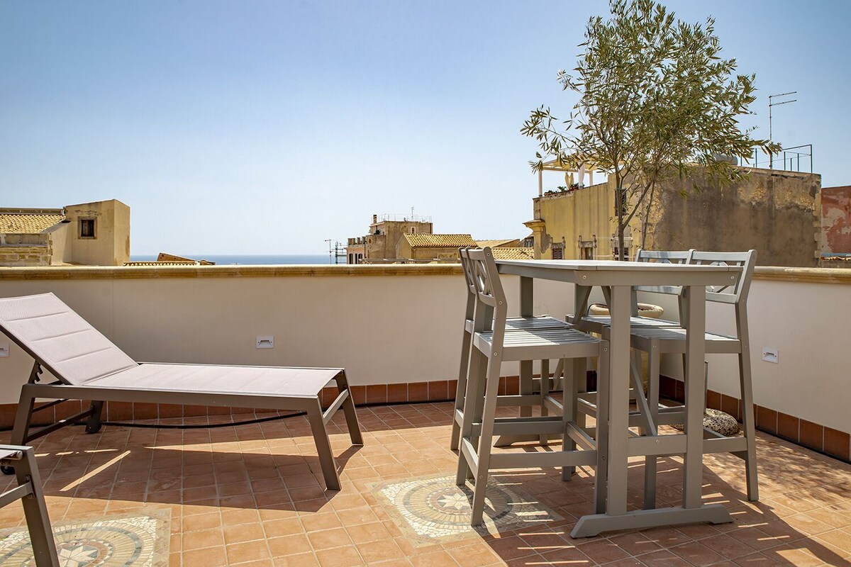 Una terrazza sul tetto con tavolo e sedie, un lettino prendisole e vista sul mare e sugli edifici.