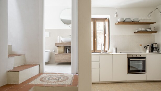 A modern kitchen with white cabinets, a built-in oven, and a coffee maker. There is a window above the sink and a door leading to another room. - La terrazza sul vicolo della Giudecca by Wonderful Italy (Syracuse)