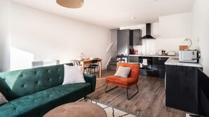 Un appartement moderne avec un canapé vert, un petit fauteuil orange, un coin cuisine et une table à manger.