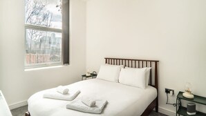 Un lit bien fait, avec du linge de lit blanc et deux serviettes enroulées, une table de chevet avec une lampe, et une fenêtre donnant sur des immeubles et des arbres sans feuilles.