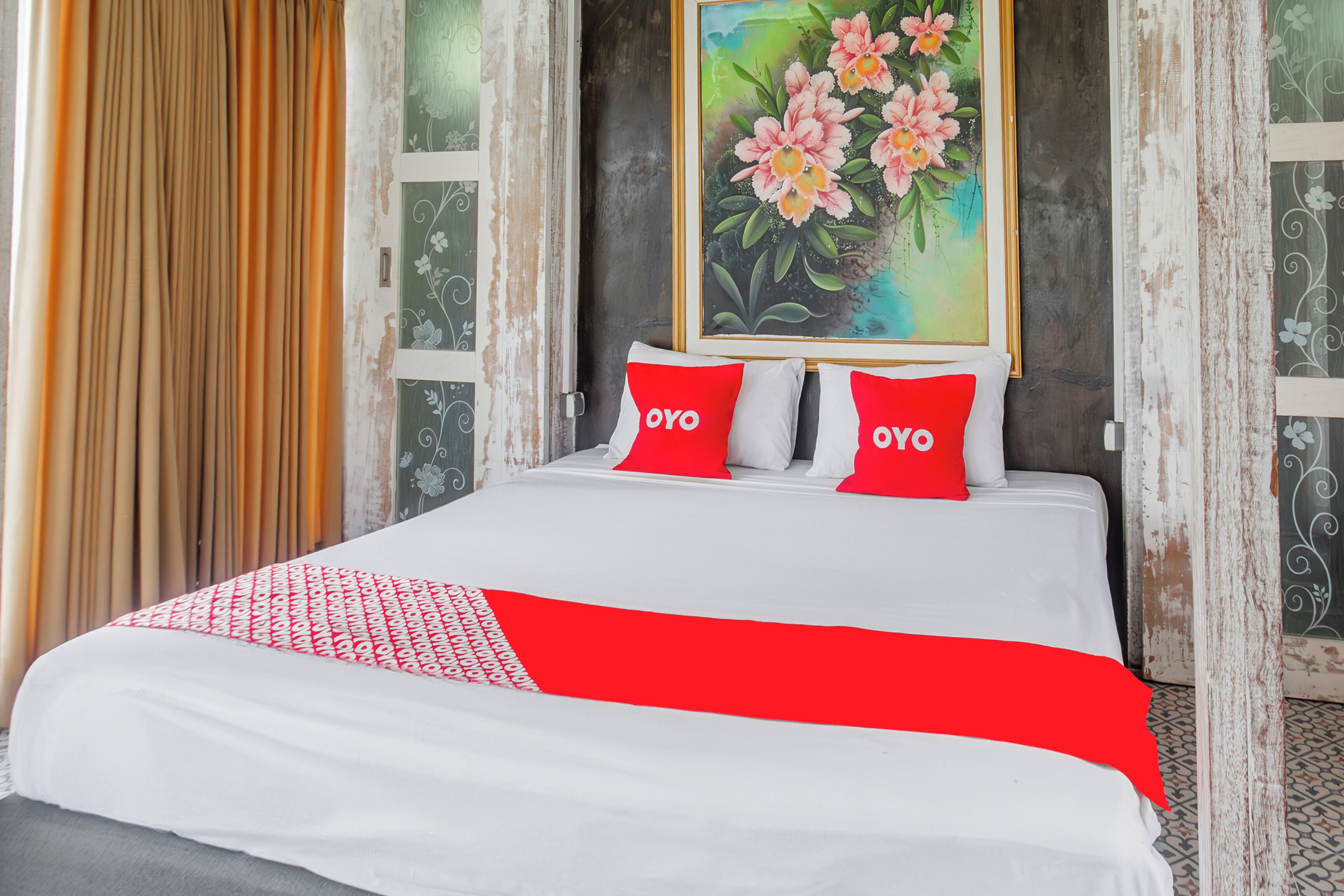A hotel room with a bed, two red pillows, a white bedspread, and a painting of flowers on the wall.