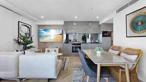 A modern living room with a sofa, dining table, and chairs, a kitchen in the background, and a beach-themed wall mural.