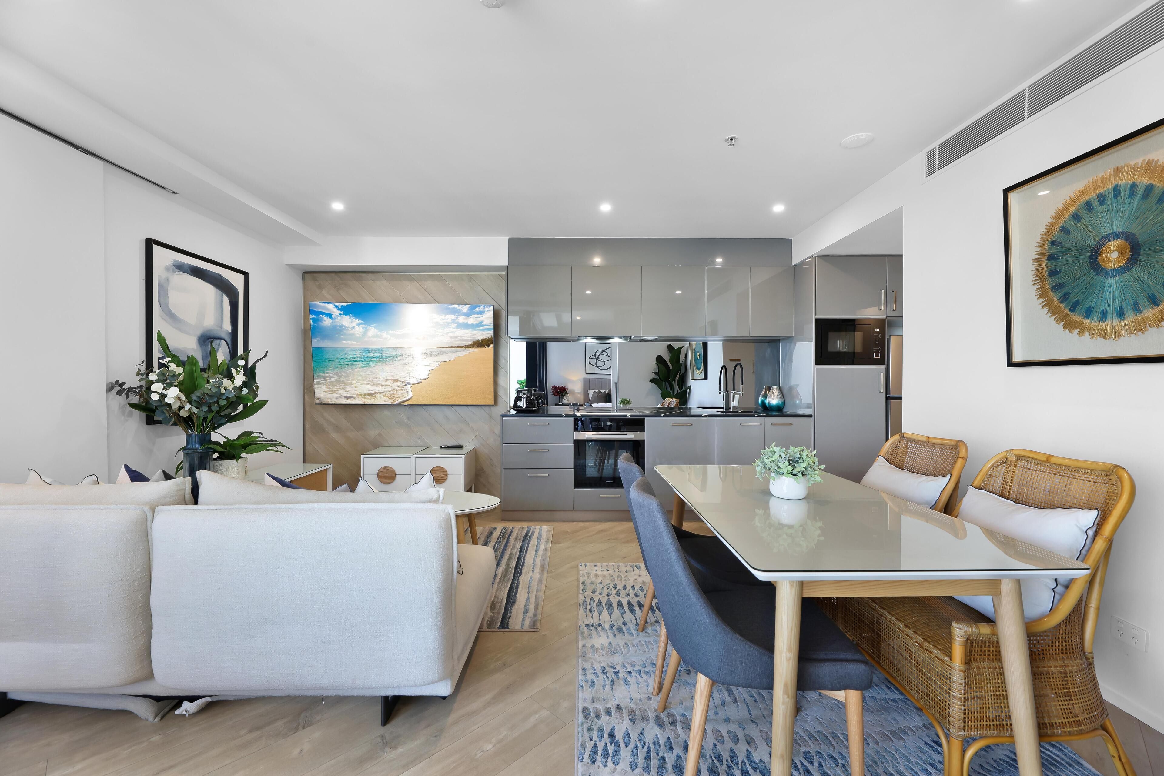 A modern living room with a sofa, dining table, and chairs, a kitchen in the background, and a beach-themed wall mural.