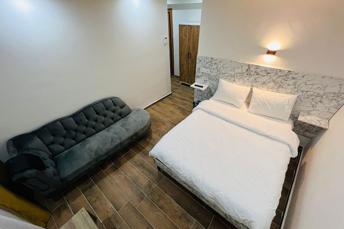 A bedroom with a bed, a grey sofa, wooden flooring, and a marble headboard.