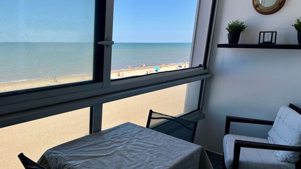 A room with a large window overlooking a beach, a table with a white tablecloth, a white chair, and a small shelf with plants. - La Land, Appartement Traversant vue mer (Courseulles-sur-Mer)