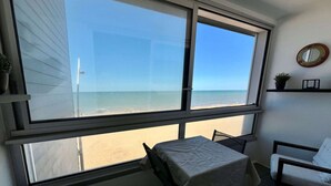 A room with a large window overlooking a beach, a table with a white tablecloth, and a chair. - La Land, Appartement Traversant vue mer (Courseulles-sur-Mer)