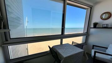 Une pièce avec une grande fenêtre donnant sur une plage, une table recouverte d’une nappe blanche et une chaise.