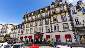 Un hotel de varios pisos con un estilo arquitectónico europeo clásico, que presenta una entrada central con toldo rojo y un balcón en el segundo piso.