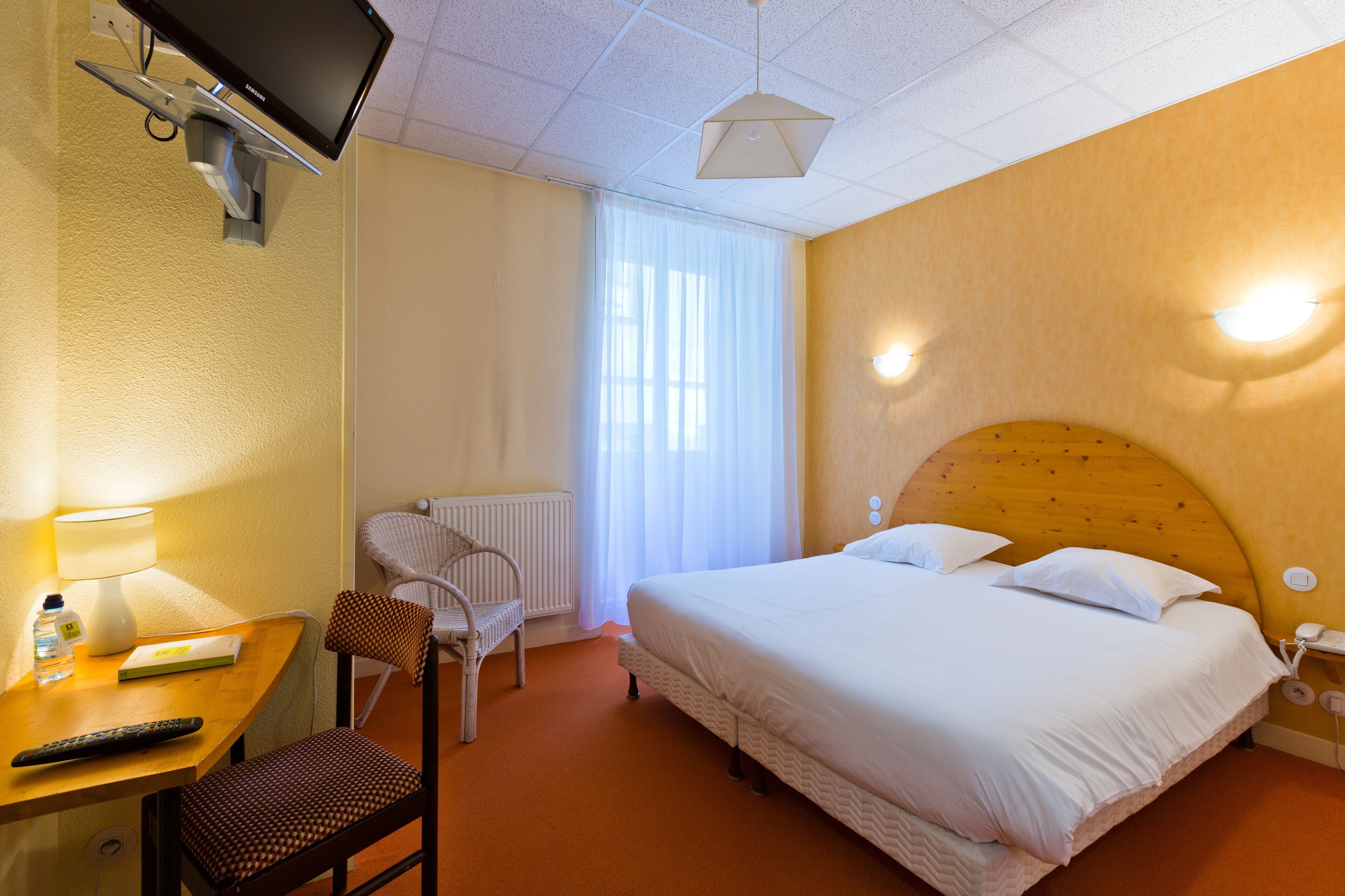 Ein Hotelzimmer mit Bett, Schreibtisch mit Lampe, Stuhl, Fernseher und einem Fenster mit Vorhängen.