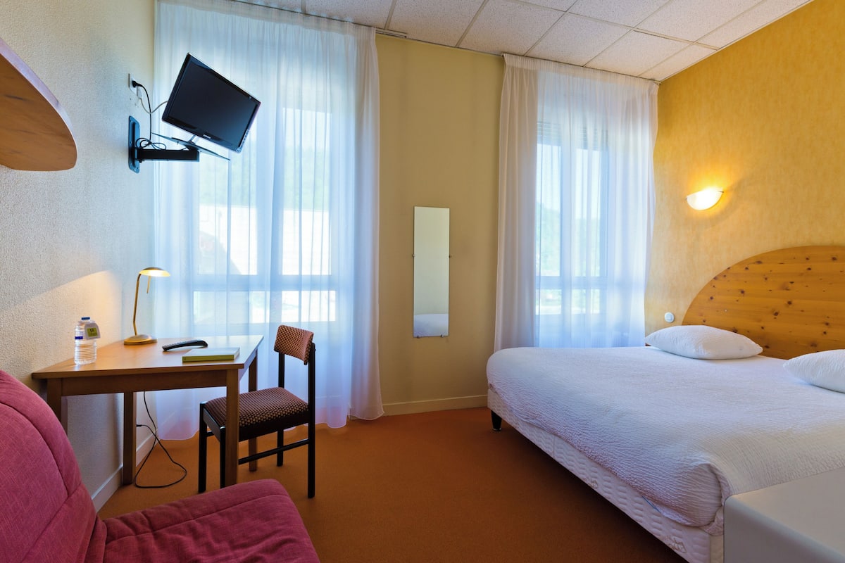 A hotel room with a bed, a desk with a chair, a television mounted on the wall, and a window with curtains.