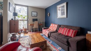 Un salon avec un canapé marron, une table basse en bois, un fauteuil rouge, un mur bleu et une fenêtre donnant sur des immeubles.