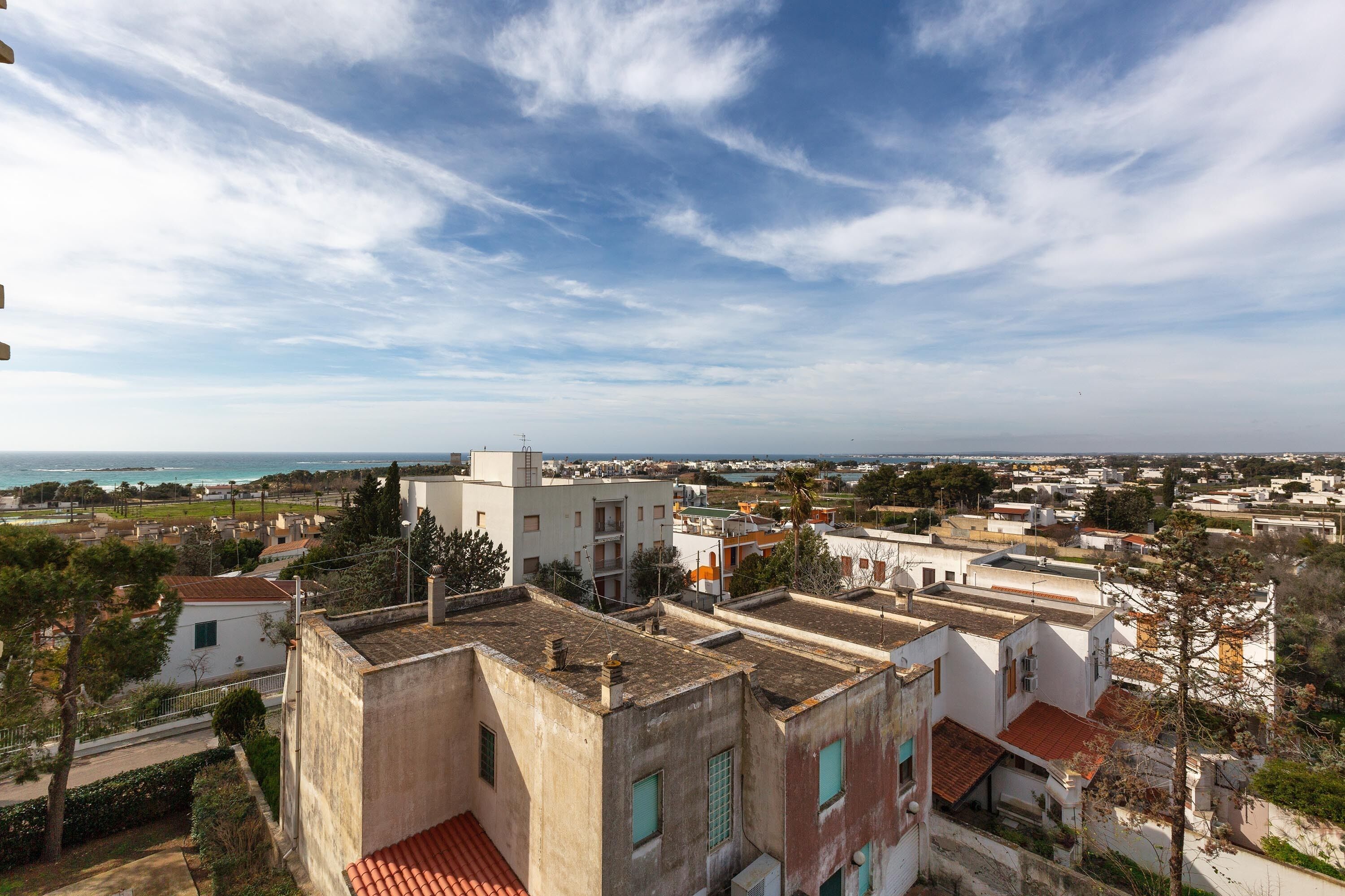 A residential area with multiple buildings, some with red-tiled roofs, and a clear view of the coastline in the distance.