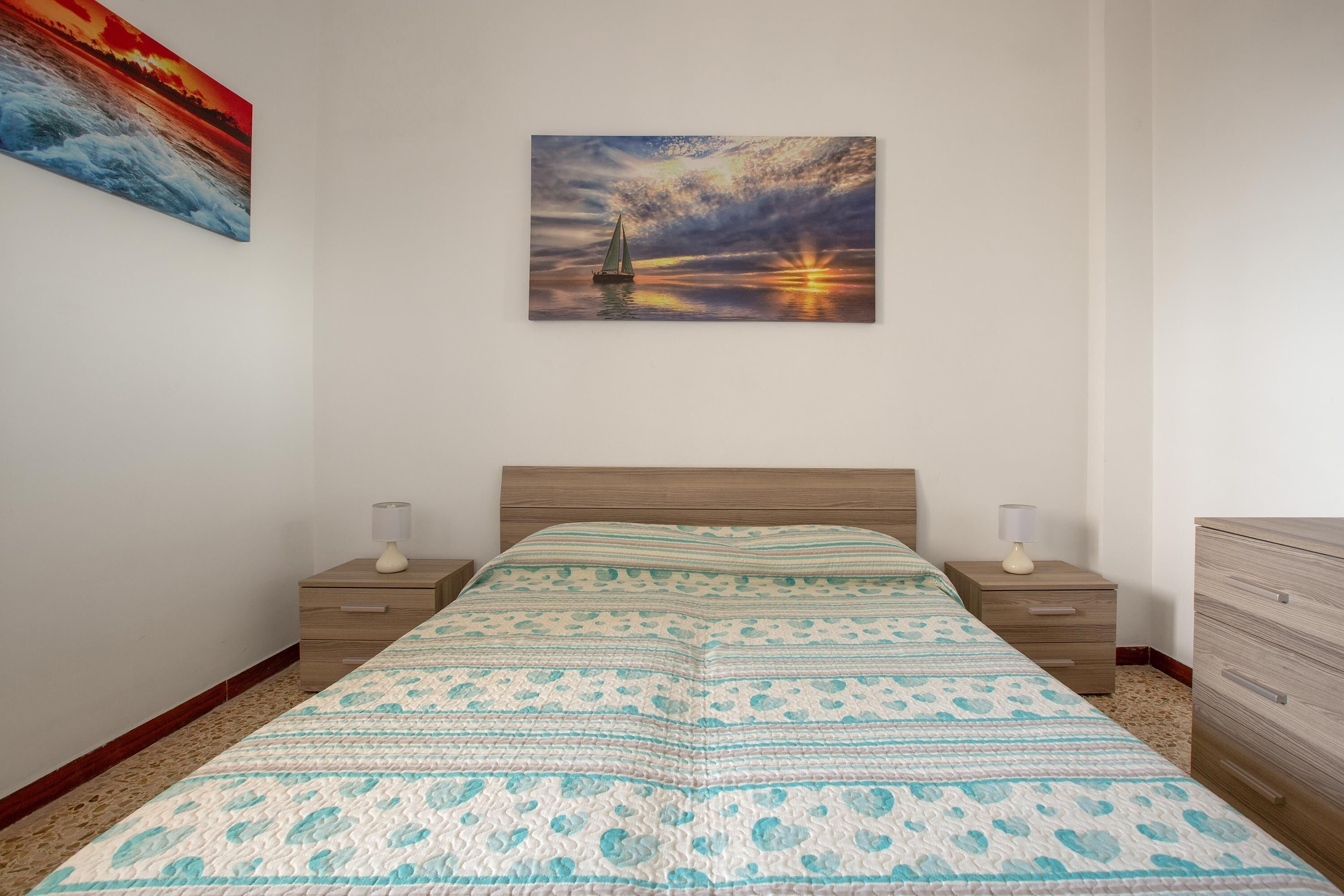 A bedroom with a bed, two bedside tables, a nightstand, a dresser, and a painting of a sunset over the ocean.