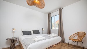 A modern bedroom with a large bed, a bedside table with a lamp, a wicker chair, and a window with curtains. - Cozy Retreat - By Wehost (Funchal)