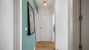 A hallway with a wooden floor, white walls, and a green accent wall. There is a framed picture on the green wall and a door at the end of the hallway. - Cozy Retreat - By Wehost (Funchal)