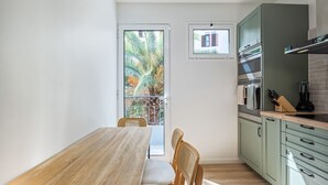 A compact kitchen with a wooden table and chairs, a small window, and a view of palm trees outside. - Cozy Retreat - By Wehost (Funchal)
