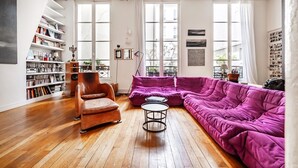 Ein modernes Wohnzimmer mit Holzboden, einem violetten Sofa, einem braunen Sessel und einem Bücherregal voller Bücher und Dekoration.