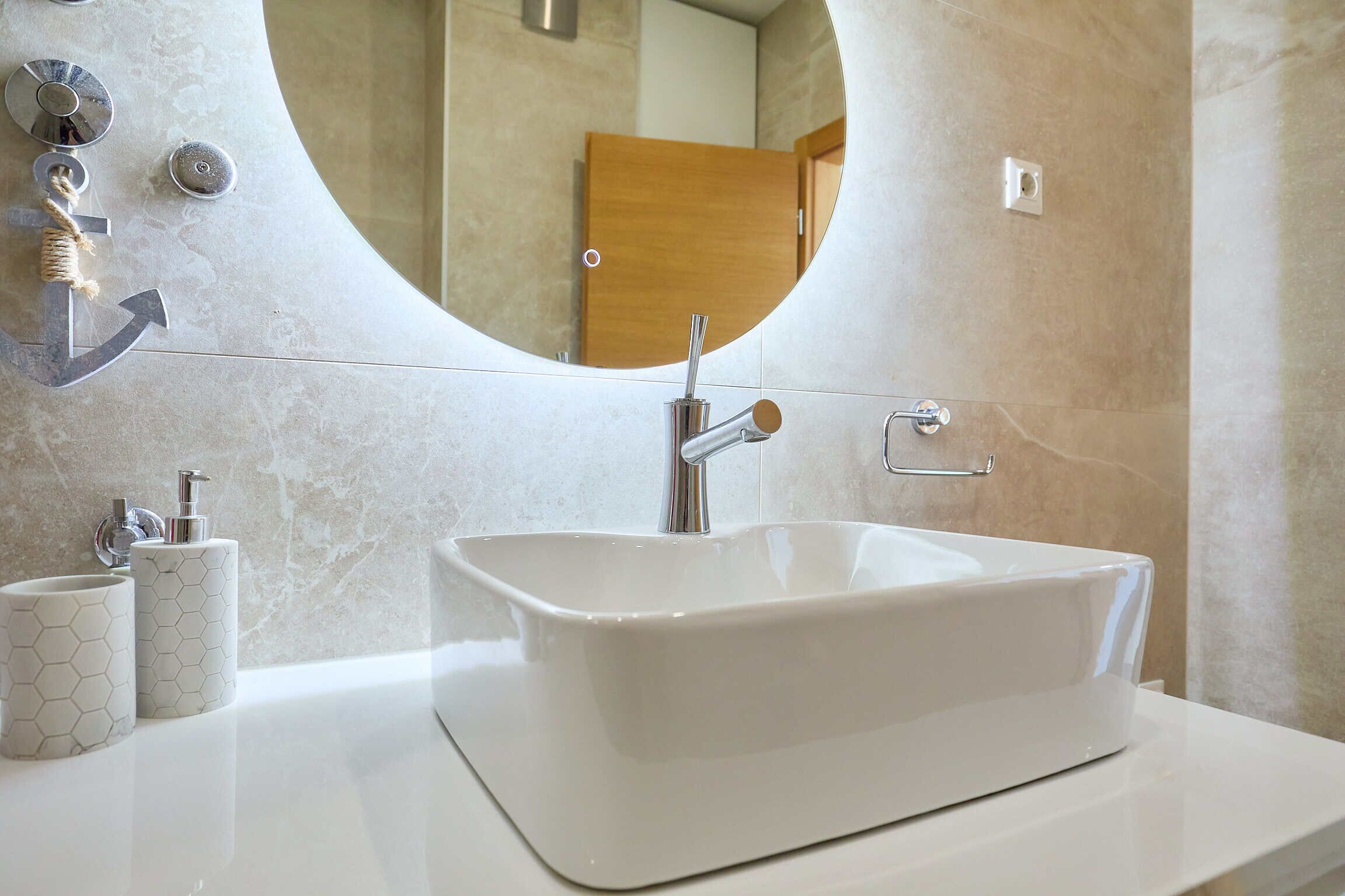 A modern bathroom with a rectangular white sink, a round mirror, and a wooden door in the background.